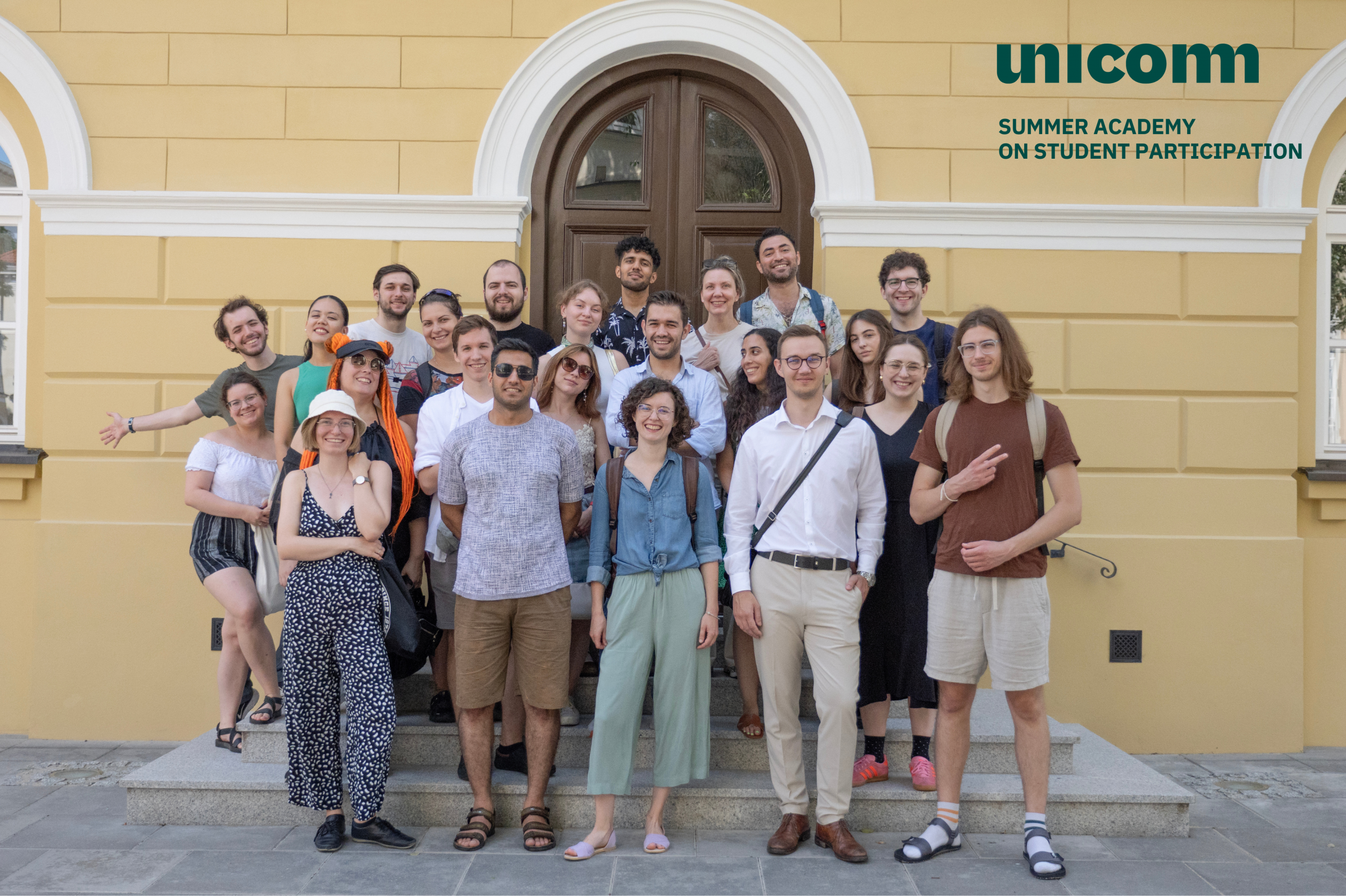 Summer Academy participants they laugh and smile in a group photo with the building at the University of Warsaw in the background